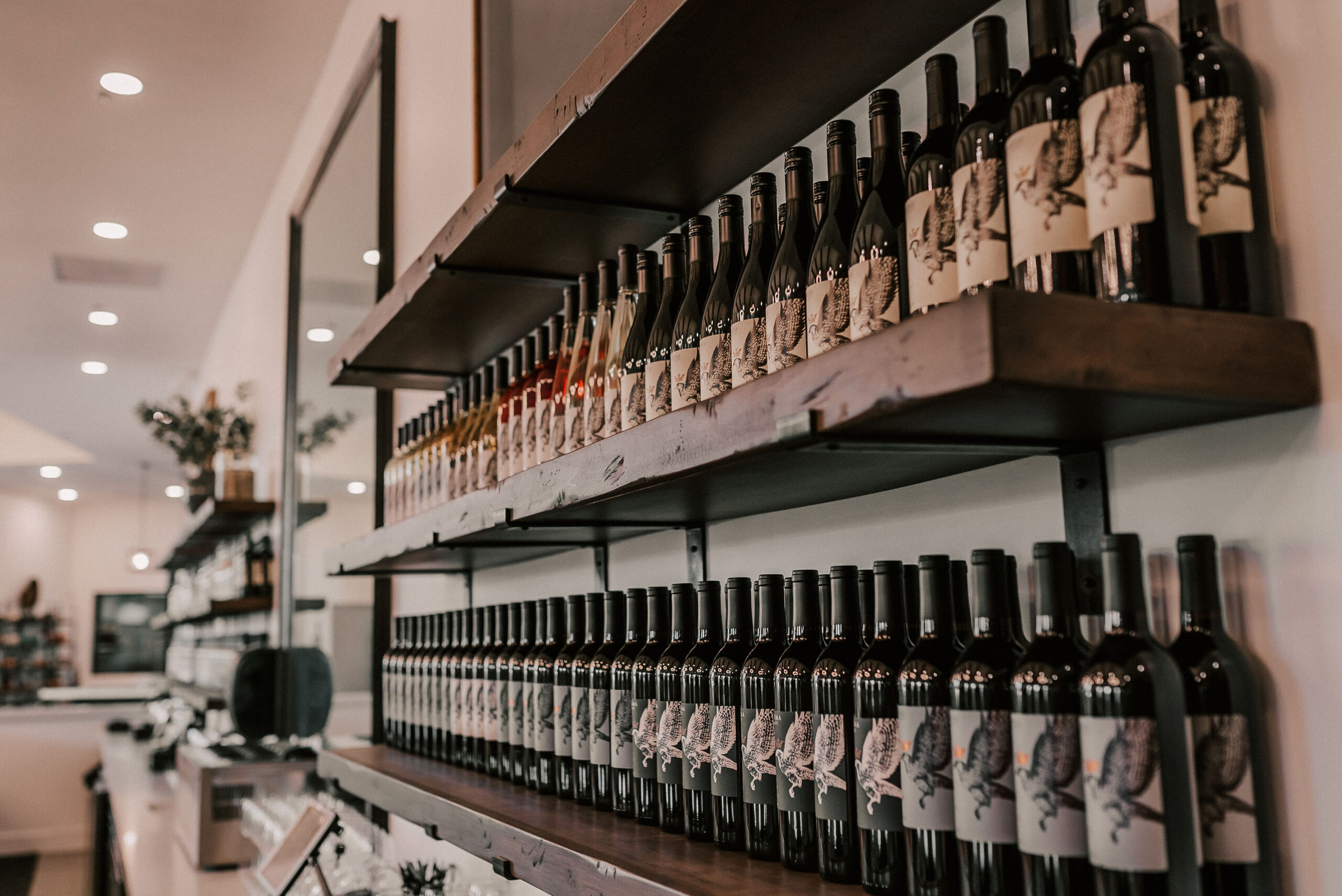 Wooden shelves displaying various bottles of wine, predominantly red, with decorative labels, inside a modern wine shop. Soft lighting enhances the ambiance.
