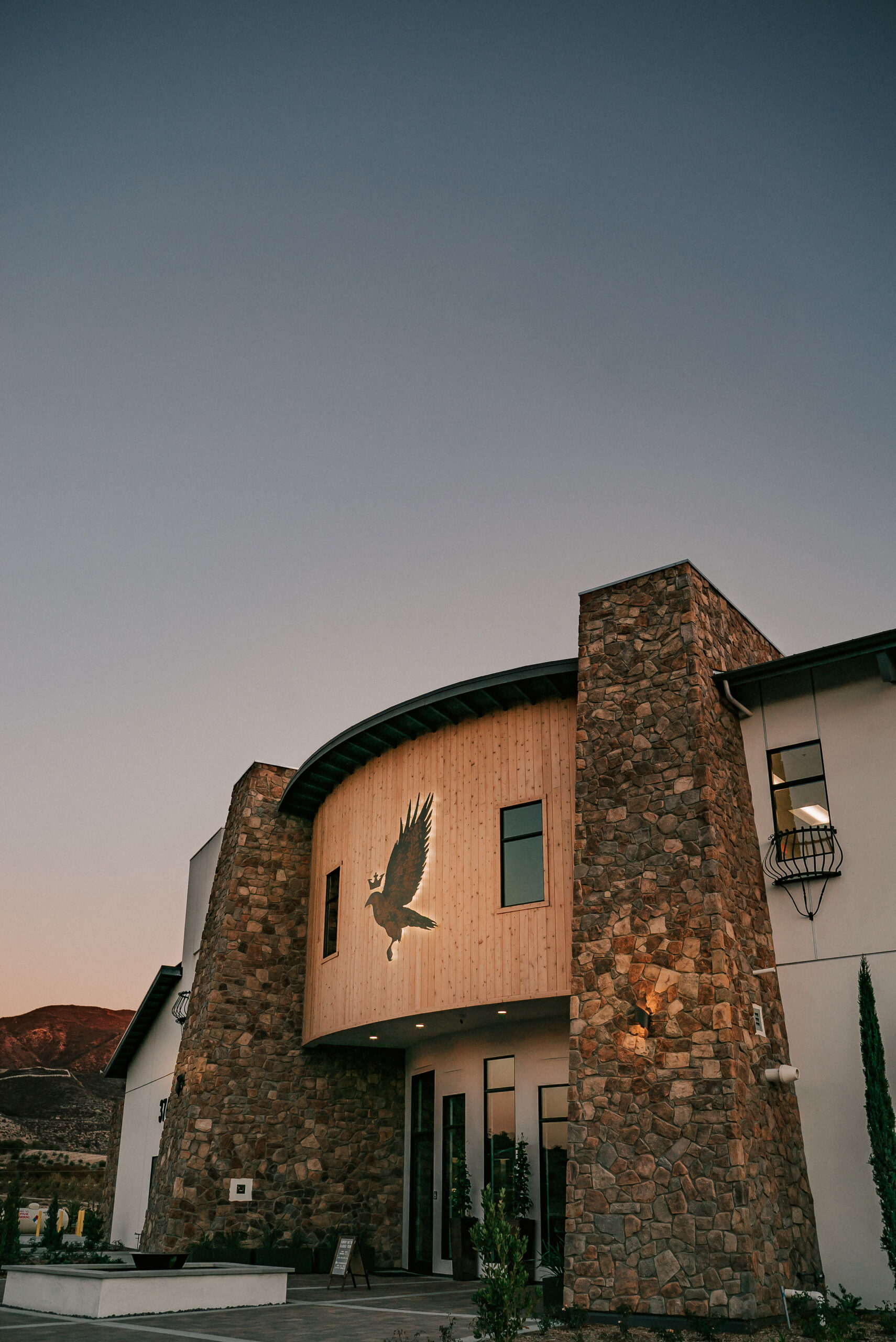 Exterior view of Altisima Winery featuring a modern stone and wood facade, with a prominent eagle emblem above the entrance. The sky is clear during dusk, enhancing the architectural details.