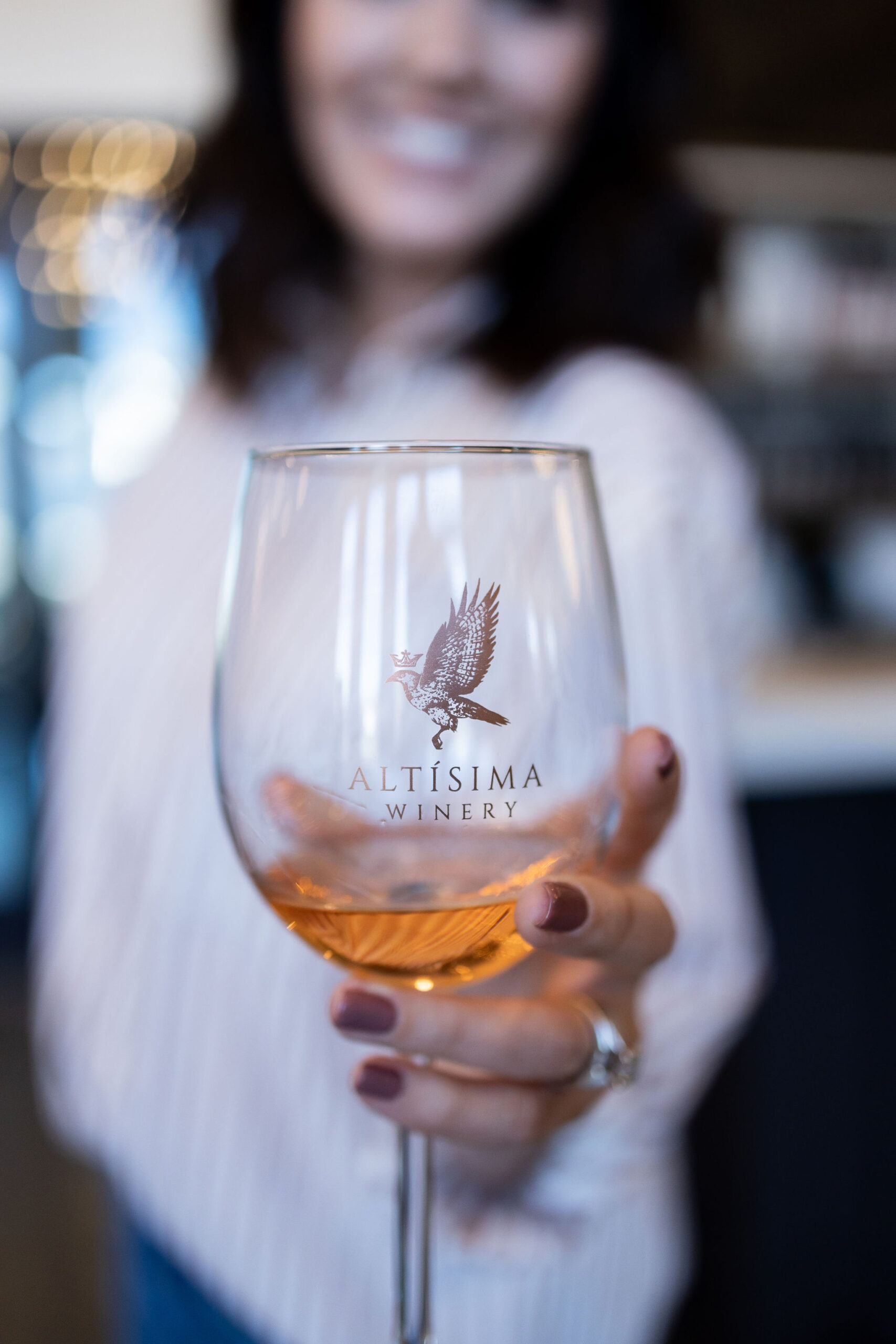 A close-up of a woman holding a clear wine glass with a rose-colored drink, featuring the logo of Altisima Winery. The background shows a softly blurred interior of a winery.