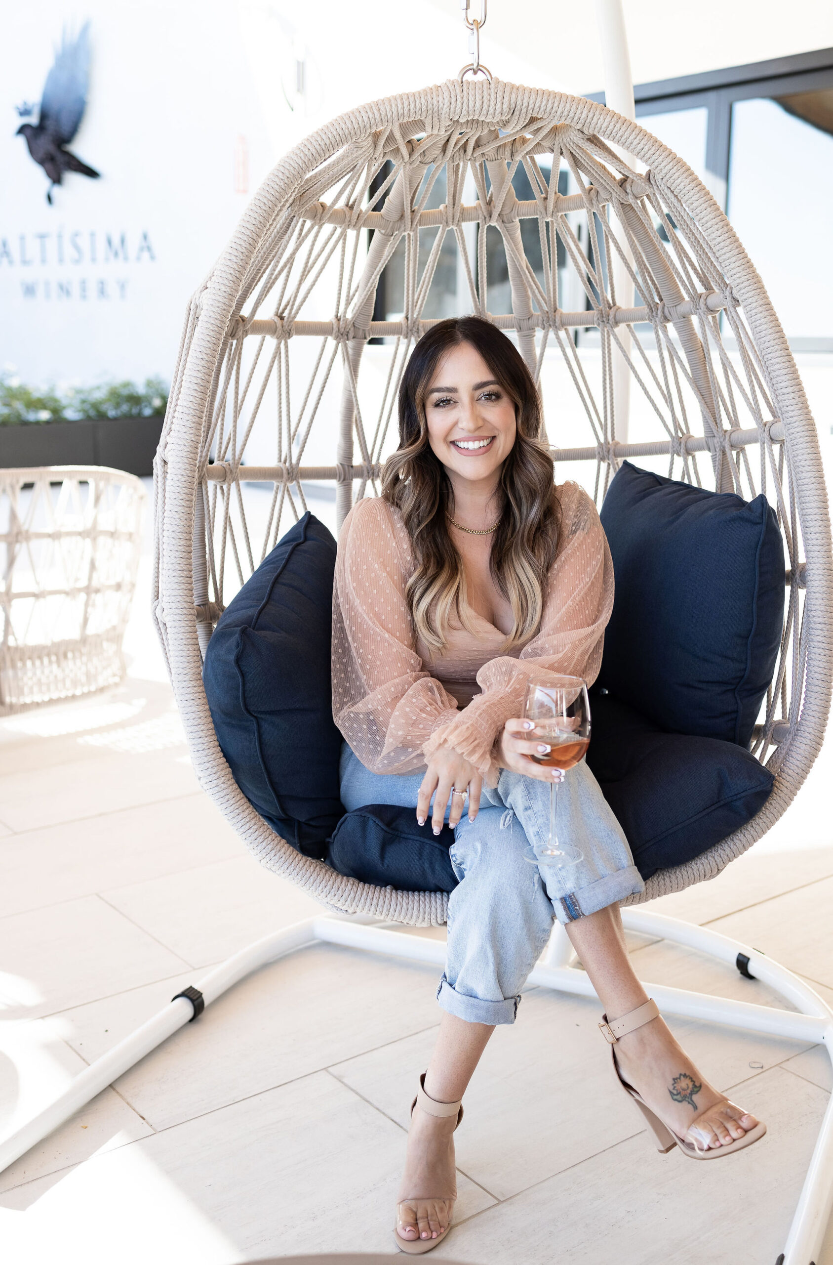 A woman sitting in a hanging chair at Altisima Winery, holding a glass of wine. She has long wavy hair, wears a sheer pink blouse, jeans, and sandals. The background features a relaxed winery setting with cushions in the chair.