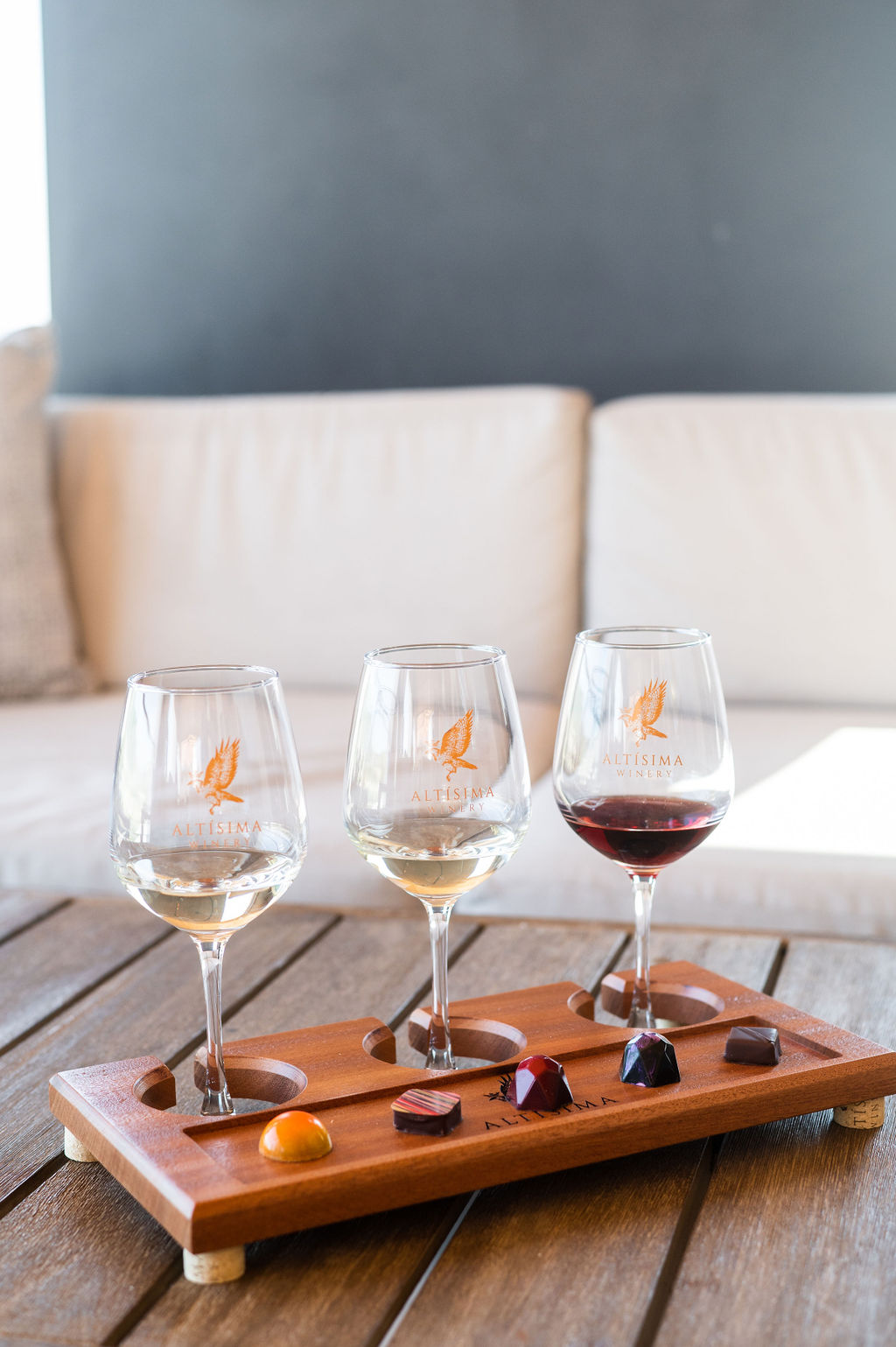 Three wine glasses holding a white wine, a rosé, and a red wine on a wooden tasting tray. The tray also holds several chocolate pieces, set against a light-colored couch and a simple background, showcasing a winery tour experience.