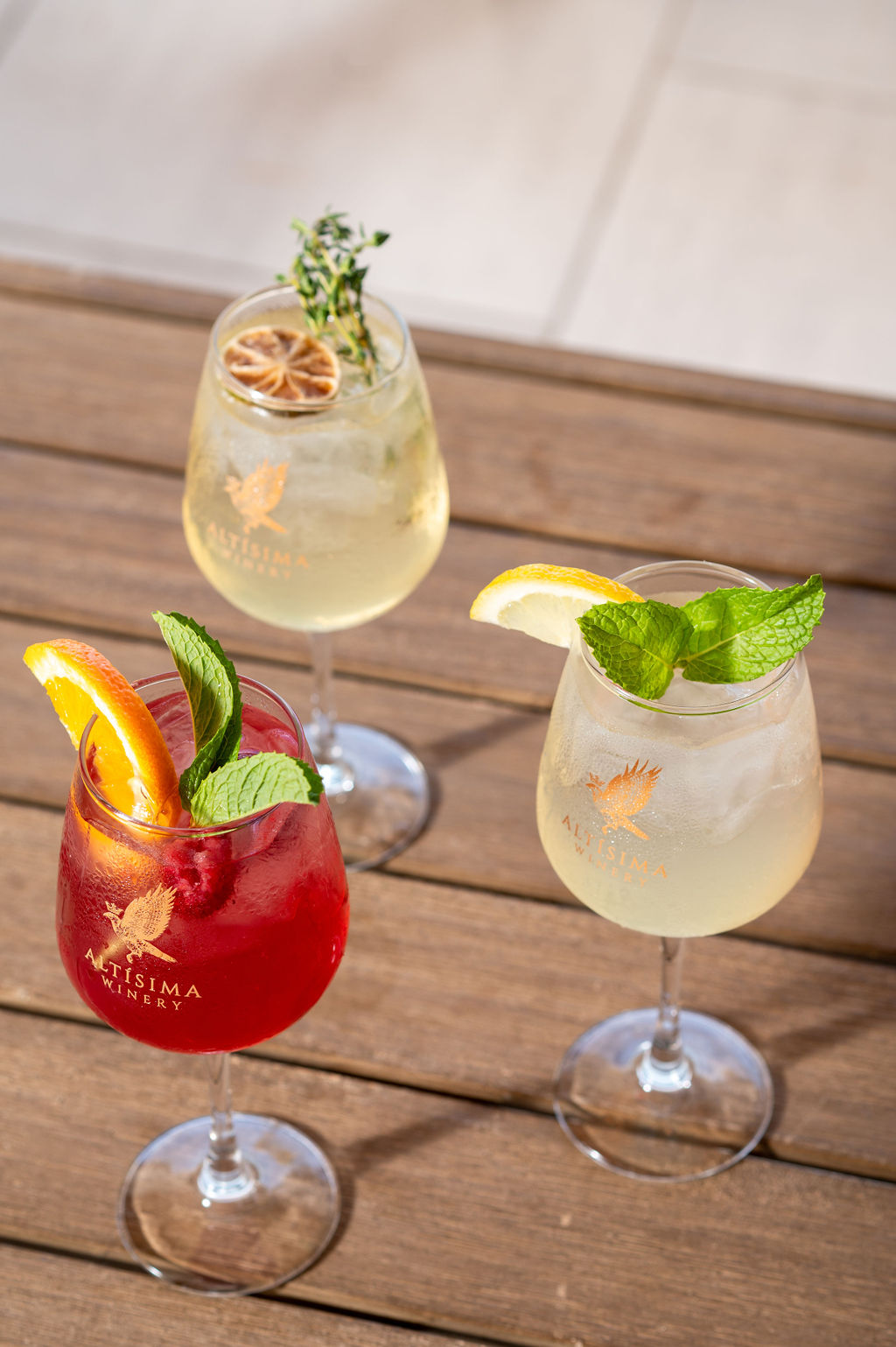 Three cocktails on a wooden surface: a red drink garnished with mint and an orange slice, a yellow drink with a slice of lemon and mint sprig, and a clear drink with a dried lime slice and thyme garnish.