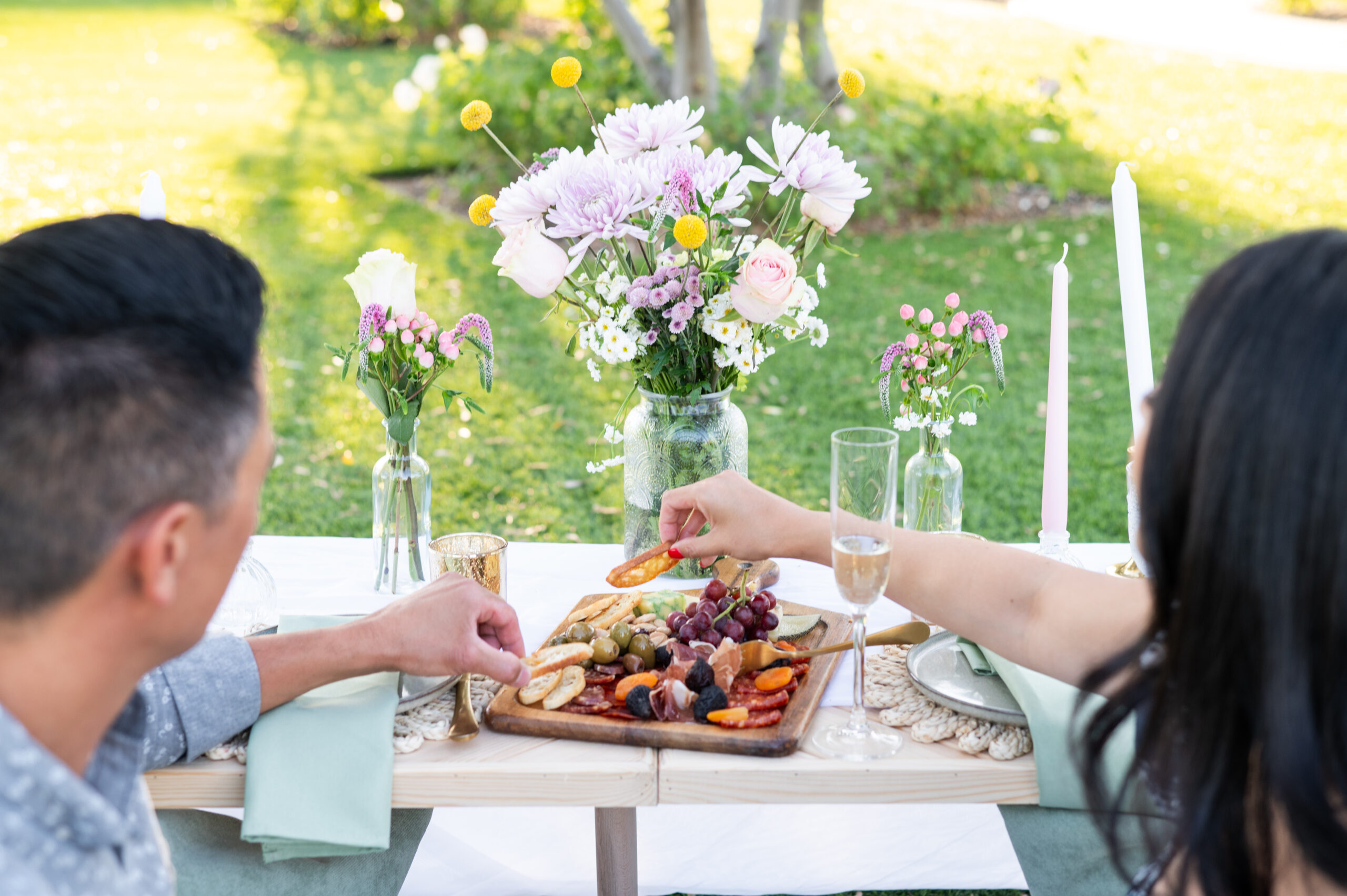 A couple enjoying a charcuterie board at an outdoor table decorated with flowers and candles. The table is set in a green area with sunlight filtering through the trees.