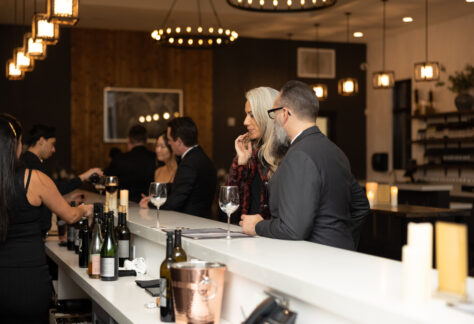 People socializing at a bar, with some individuals standing and others seated. The bar features a variety of drinks on display and is illuminated by hanging lights, creating a modern and inviting atmosphere.