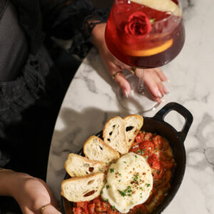 A person holds a black skillet of baked pasta topped with melted cheese and toasted bread slices, with a red cocktail on a marble table.