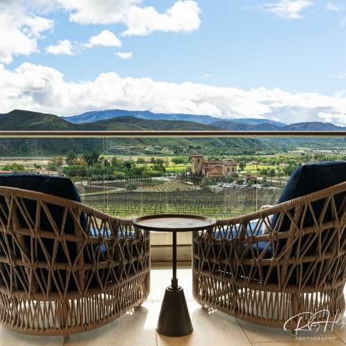 Two woven lounge chairs facing a scenic view of mountains and green valleys through a glass railing. A small round table is placed between the chairs, creating a cozy atmosphere.