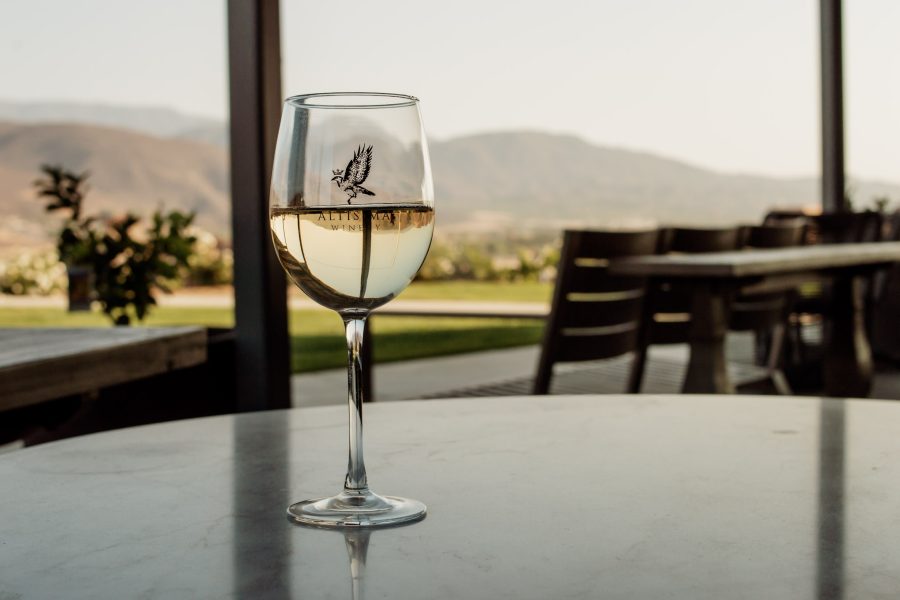 A glass of white wine with a logo, placed on a marble table. In the background, there are outdoor seating areas, greenery, and mountains under clear skies, typical of a vineyard setting.