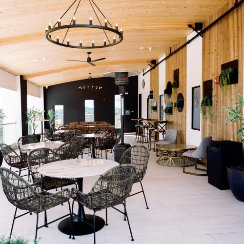 An interior view of a modern restaurant featuring a wooden ceiling, circular chandelier, and a variety of seating options including woven chairs and wooden tables. Green plants are placed around the space, with art displayed on the walls.