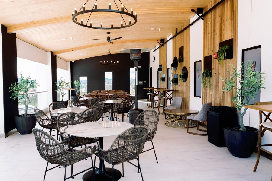 An interior view of a modern restaurant featuring a wooden ceiling, circular chandelier, and a variety of seating options including woven chairs and wooden tables. Green plants are placed around the space, with art displayed on the walls.