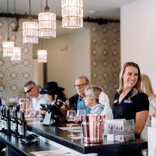 A smiling staff member serves guests at a wine tasting bar. Several patrons are seated at the counter, with wine bottles and glasses visible. The decor includes elegant light fixtures and patterned walls, creating a welcoming atmosphere for the wine club.