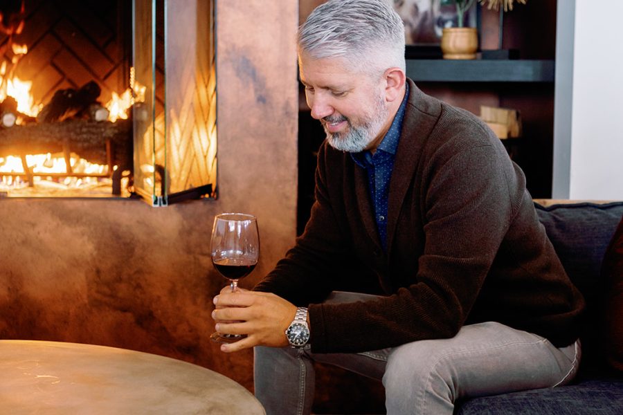 A man with gray hair sits on a dark sofa, holding a glass of wine. He wears a dark sweater over a blue shirt, smiling softly. In the background, a warm fireplace crackles against a stylish interior featuring bookshelves and decor.