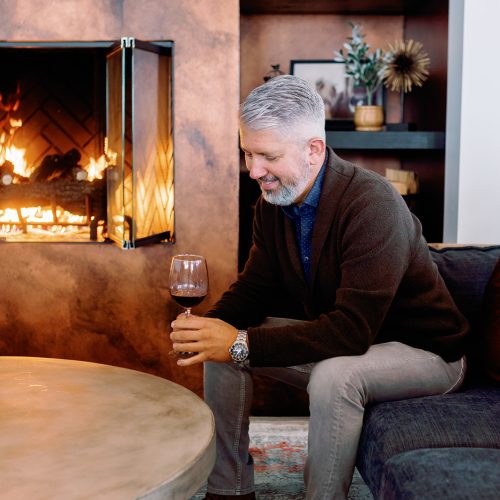A smiling man holding a wine glass while seated on a couch, with a cozy fireplace in the background. The scene features warm decor, including bookshelves and decorative elements, creating a relaxed atmosphere ideal for a wine club gathering.