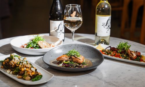 A table set with three gourmet dishes featuring cooked salmon with greens, roasted Brussels sprouts, and a vibrant vegetable salad. Two bottles of white wine and a glass of wine are placed on the table, suggesting a dining experience at Gaspar's Restaurant, known for Spanish Fusion dishes.