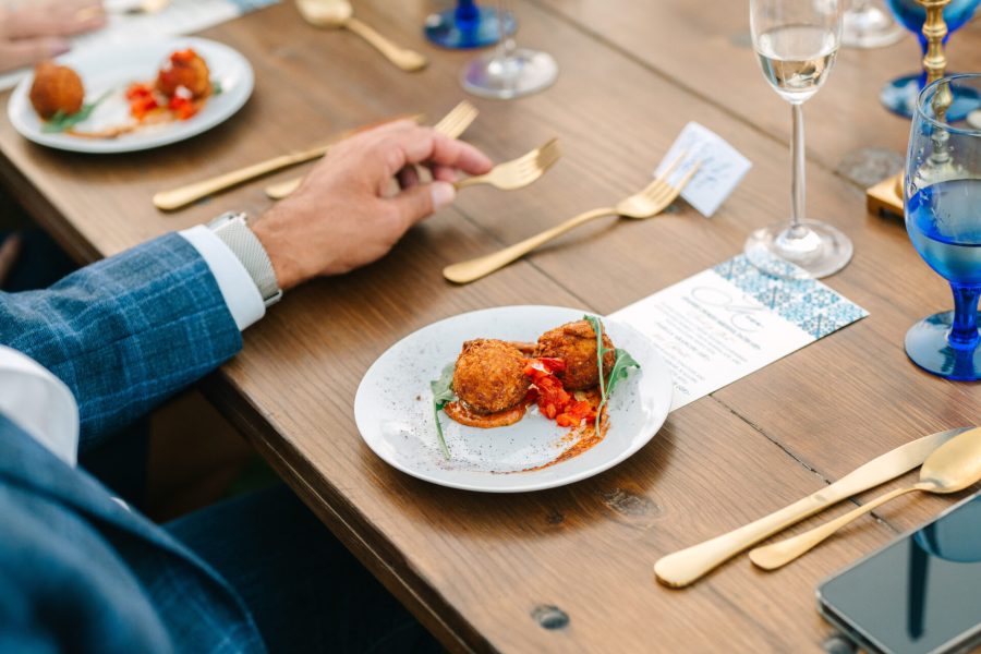 A table set with elegant place settings featuring golden utensils and blue glassware. A close-up shows a hand holding a plate with two meatballs in sauce, garnished with a leaf, alongside a place card and glassware.