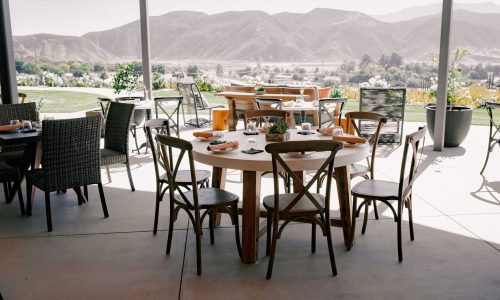 An outdoor dining area featuring wooden and wicker furniture, including tables and chairs. In the background, mountains and greenery are visible under a clear sky.