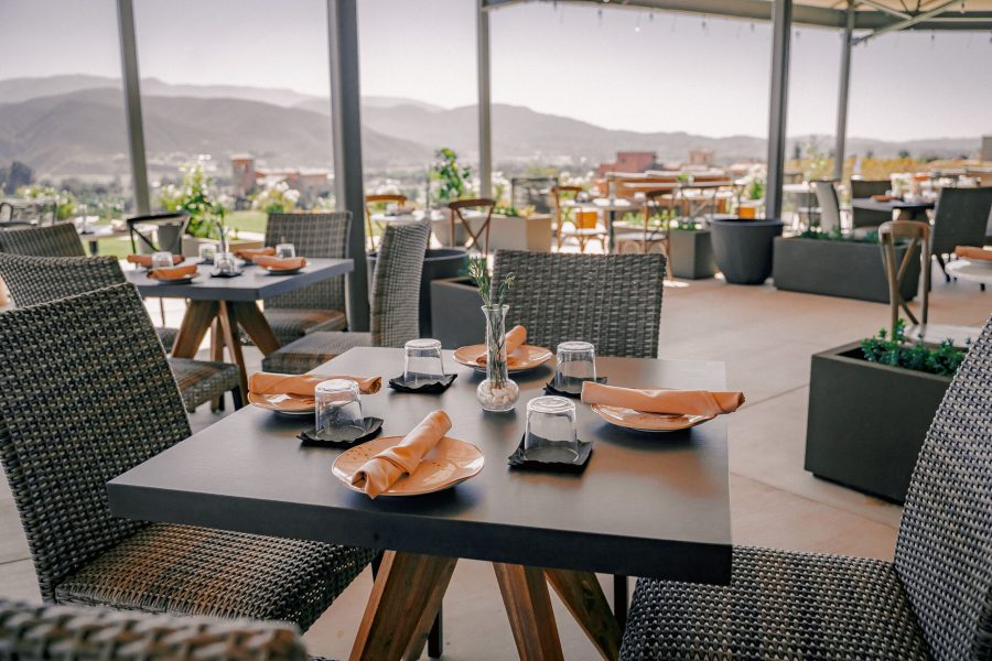 An outdoor dining area featuring wicker tables and chairs with brown napkins placed on black plates. Clear glass cups are on the tables, with views of distant mountains and greenery visible in the background.