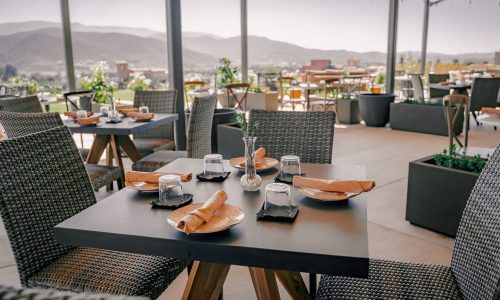 A restaurant patio with tables set for dining, featuring plates, utensils, and glasses. The background shows a scenic view of mountains and distant buildings under a clear sky.