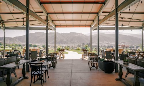 An open dining area with wooden tables and chairs, surrounded by mountains in the background. The space is covered, with string lights hanging from the ceiling, creating a relaxed outdoor dining atmosphere.
