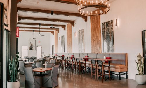 An indoor restaurant featuring modern decor, with wooden beams and light fixtures. Seating includes upholstered booths and wooden tables. Large windows allow natural light, and plants are placed throughout the space.