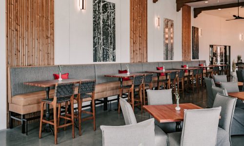 Interior of a modern restaurant featuring wooden panel walls and seating. There are high tables with stools and booths with cushions, creating a spacious dining atmosphere. Artworks adorn the walls, enhancing the decor.