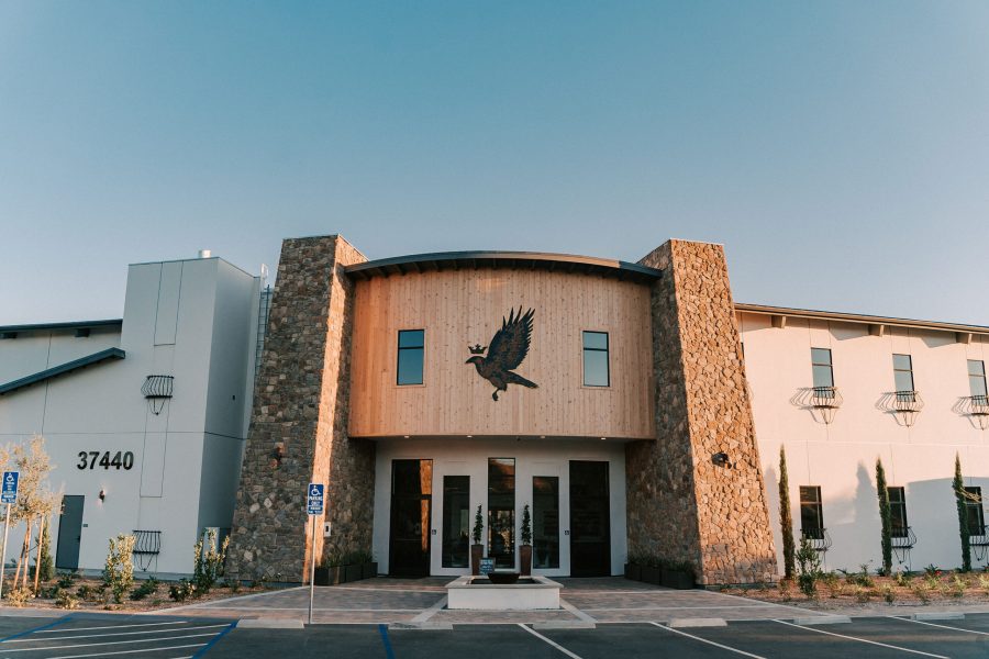 A modern two-story building with a curved wooden entrance, featuring a large eagle emblem above the doors. The facade includes stone columns and parking spaces in front. Clear blue sky above.
