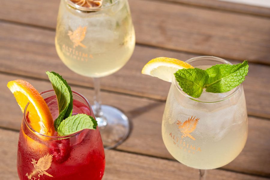 Three cocktails on a wooden surface: a red drink garnished with mint and an orange slice, a yellow drink with a slice of lemon and mint sprig, and a clear drink with a dried lime slice and thyme garnish.