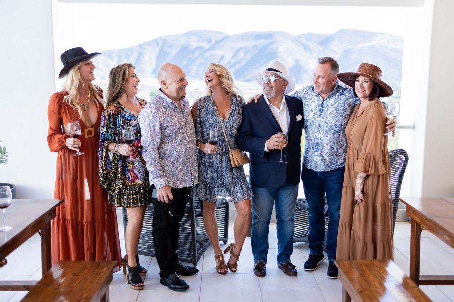 A group of seven adults dressed in stylish outfits, smiling and interacting in a bright indoor setting with mountains visible through a window. They are holding drinks, with wooden tables in the foreground.
