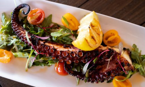 A grilled octopus tentacle served on a bed of arugula, garnished with lemon wedges and cherry tomatoes on a white rectangular plate.