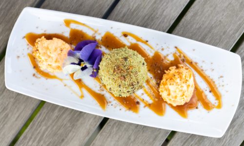A serving plate with three scoops of ice cream: one orange, one green coated in nuts, and one peach, drizzled with caramel sauce, and garnished with a purple flower. Ideal for showcasing desserts at Gaspar's Restaurant, known for Spanish fusion dishes in Temecula.