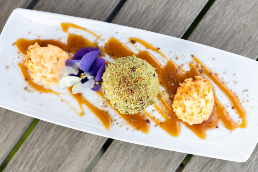 A serving plate with three scoops of ice cream: one orange, one green coated in nuts, and one peach, drizzled with caramel sauce, and garnished with a purple flower. Ideal for showcasing desserts at Gaspar's Restaurant, known for Spanish fusion dishes in Temecula.