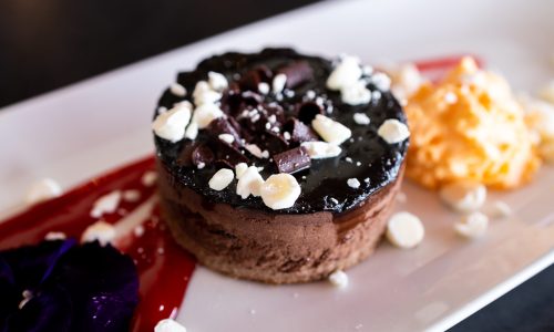 A chocolate dessert with a glossy top, chocolate shavings, and white sprinkles, served on a white plate. There is a red sauce and a scoop of orange dessert next to it, with edible flowers adding color.