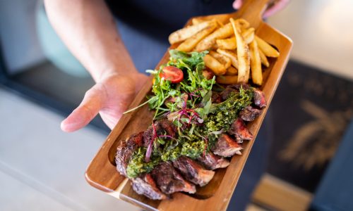 A chef holding a wooden platter featuring sliced steak topped with green sauce, accompanied by a small salad and golden French fries. The setting is contemporary, with a hint of elegance, suitable for Gaspar's Restaurant, known for Spanish Food and Spanish Fusion.