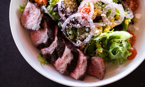 A white bowl filled with sliced grilled steak served alongside a fresh salad of leafy greens, cherry tomatoes, corn, red onions, and cheese. The dish showcases the Spanish fusion flavors of Gaspar's Restaurant.