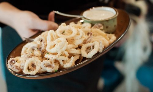 A person holding a platter of crispy calamari rings alongside a small bowl of dipping sauce, wearing a black apron. Soft focus on the background people and setting.