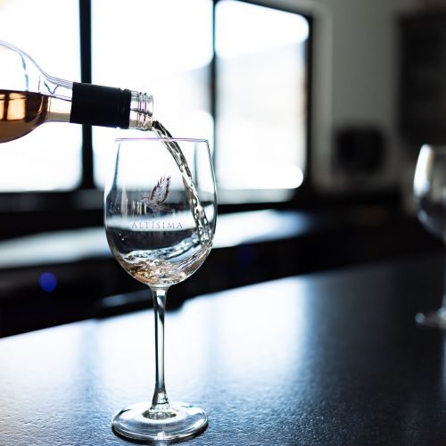 A person pouring rosé wine from a bottle into a glass with the winery logo, set on a black countertop. Two additional empty wine glasses are visible in the background, along with a large window letting in natural light.