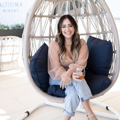 A woman sitting in a hanging chair at Altisima Winery, holding a glass of wine. She has long wavy hair, wears a sheer pink blouse, jeans, and sandals. The background features a relaxed winery setting with cushions in the chair.